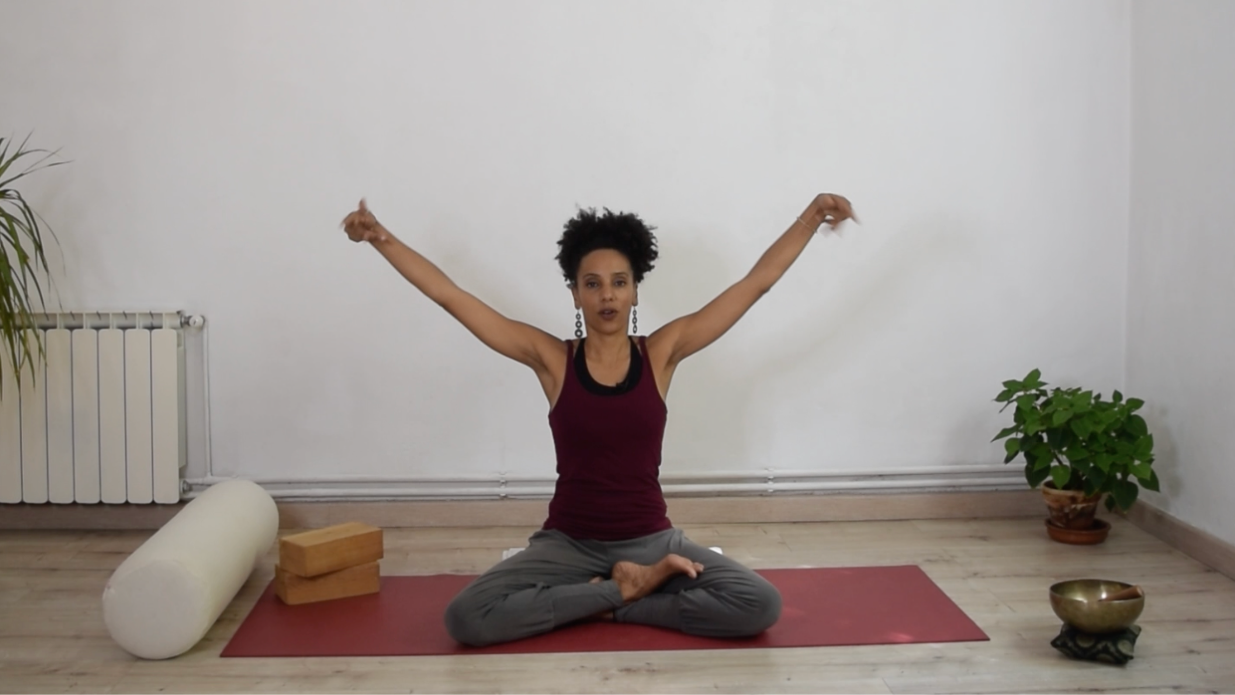 A person sitting upright on a yoga mat, making soft fists and circling their wrists while raising their arms overhead.